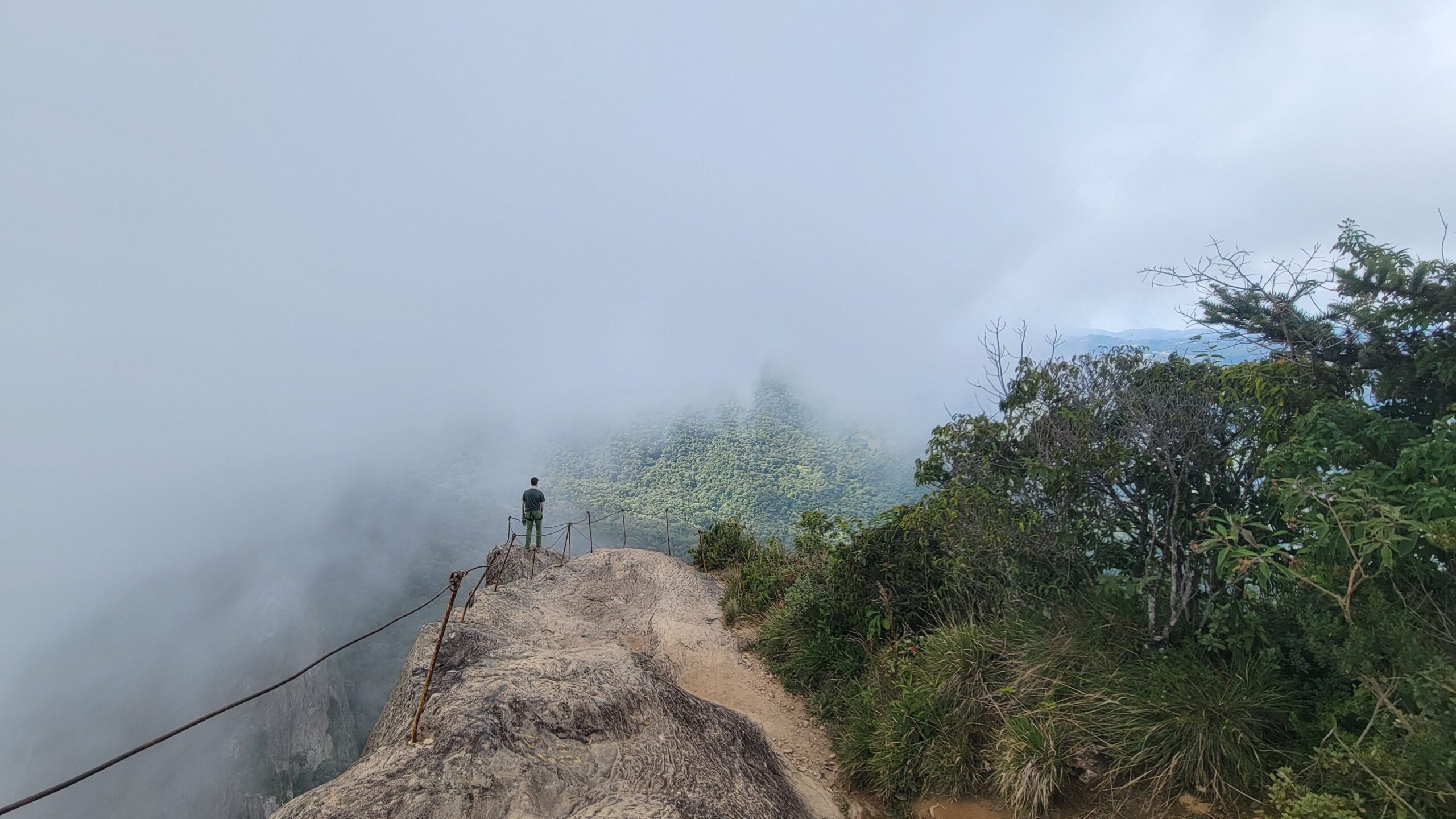 a photo of a man standing on the top of a mountain hiking trail with clouds rolling in from the left to the right.